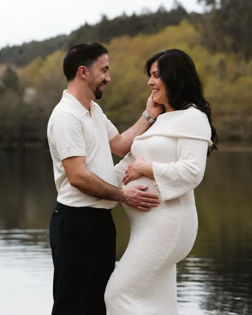 Um casal à espera de bebé, junto à água. Ele toca-lhe na cara e ela segura a barriga.