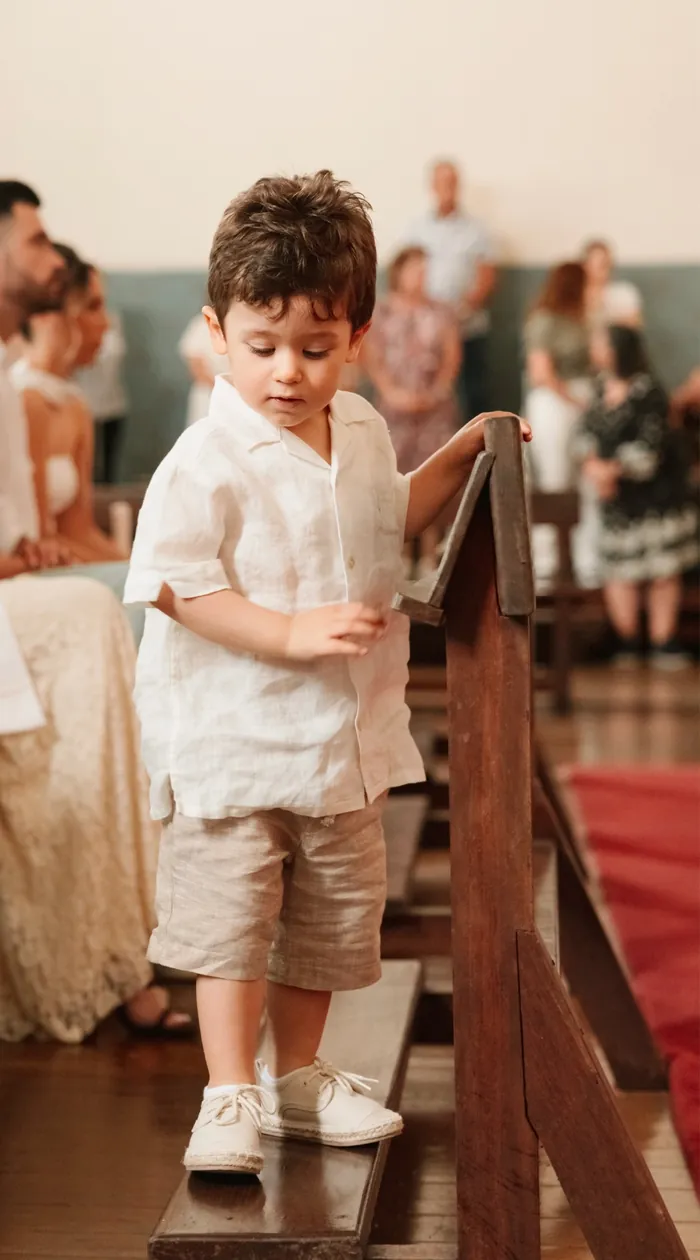 Menino de camisa de linho, de pé num banco da igreja, com os convidados ao fundo.