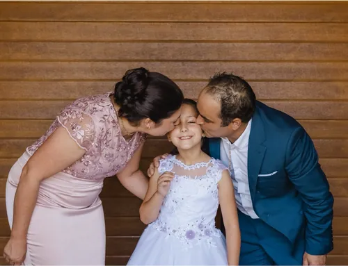 Menina com o vestido da comunhão, a receber um beijo do pai e da mãe ao mesmo tempo.