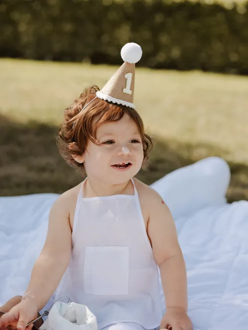 Menino de um ano com chapéu de festa e bolo no nariz, numa sessão ao ar livre.